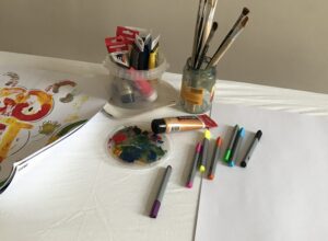 Auf einem Tisch mit weisser Decke liegen eine Auswahl an Stiften und ein Blatt weisses Papier, sowie ein Behälter mit Farbtuben und ein Glas mit Pinseln. am Linken Bildrand ist ein Teil eines bunt bemalten Papiers zu sehen.